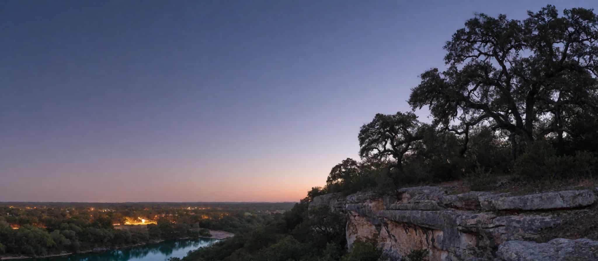A limestone bluff and live oaks above a river at twilight, with small-town lights in the distance.